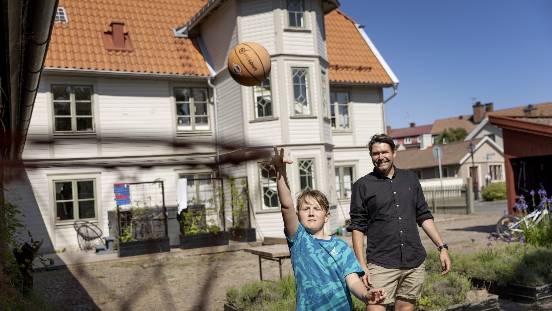Far och son spelar basket i trädgården, med en stor trävilla i bakgrunden.