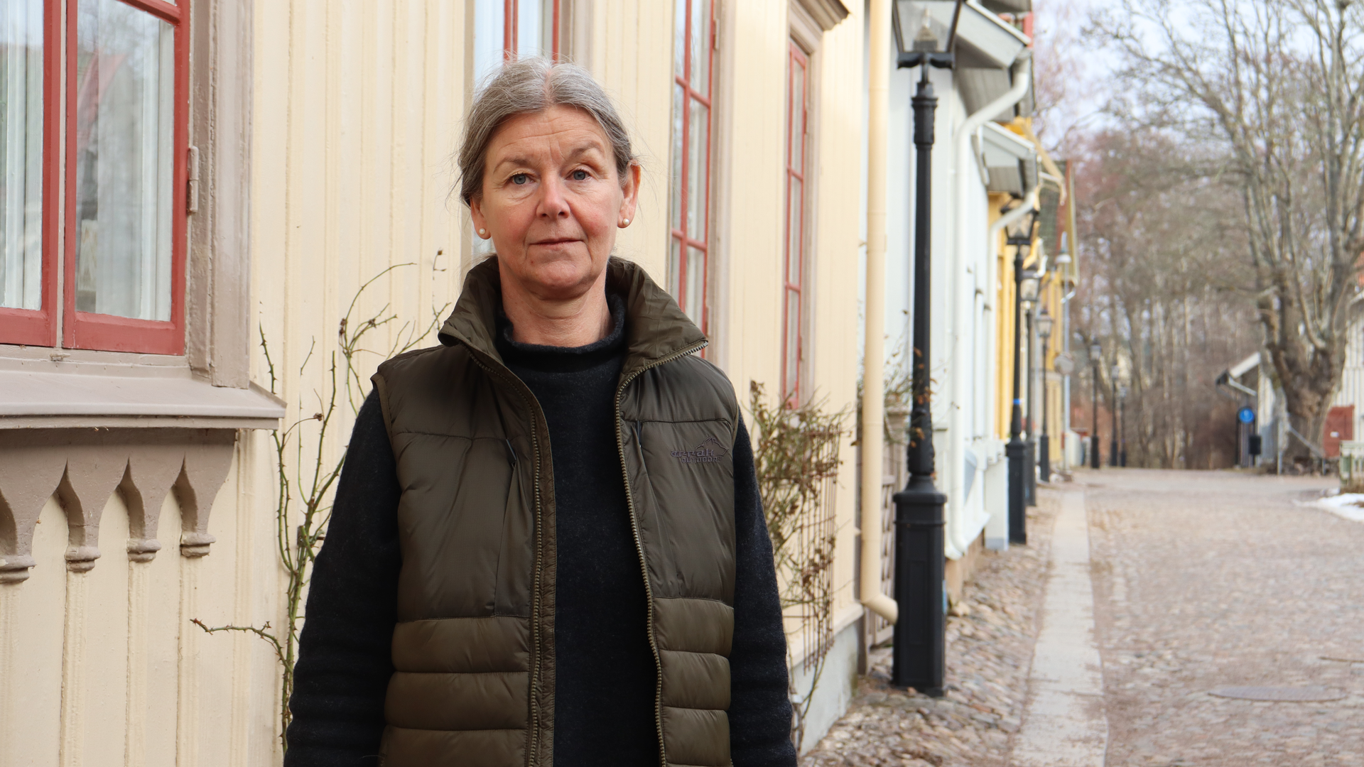 Maria Berg, samhällsbyggnadschef, står vid en trävilla på en kullerstensgata.