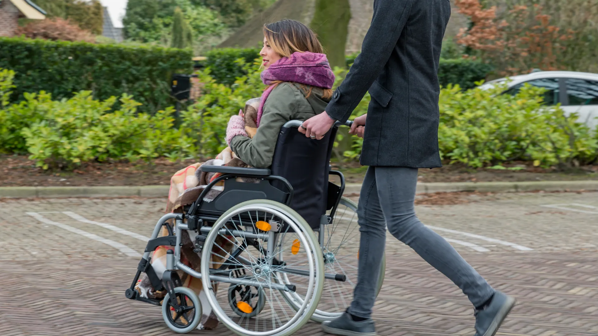 En man skjutsar en kvinna i rullstol över en parkering.