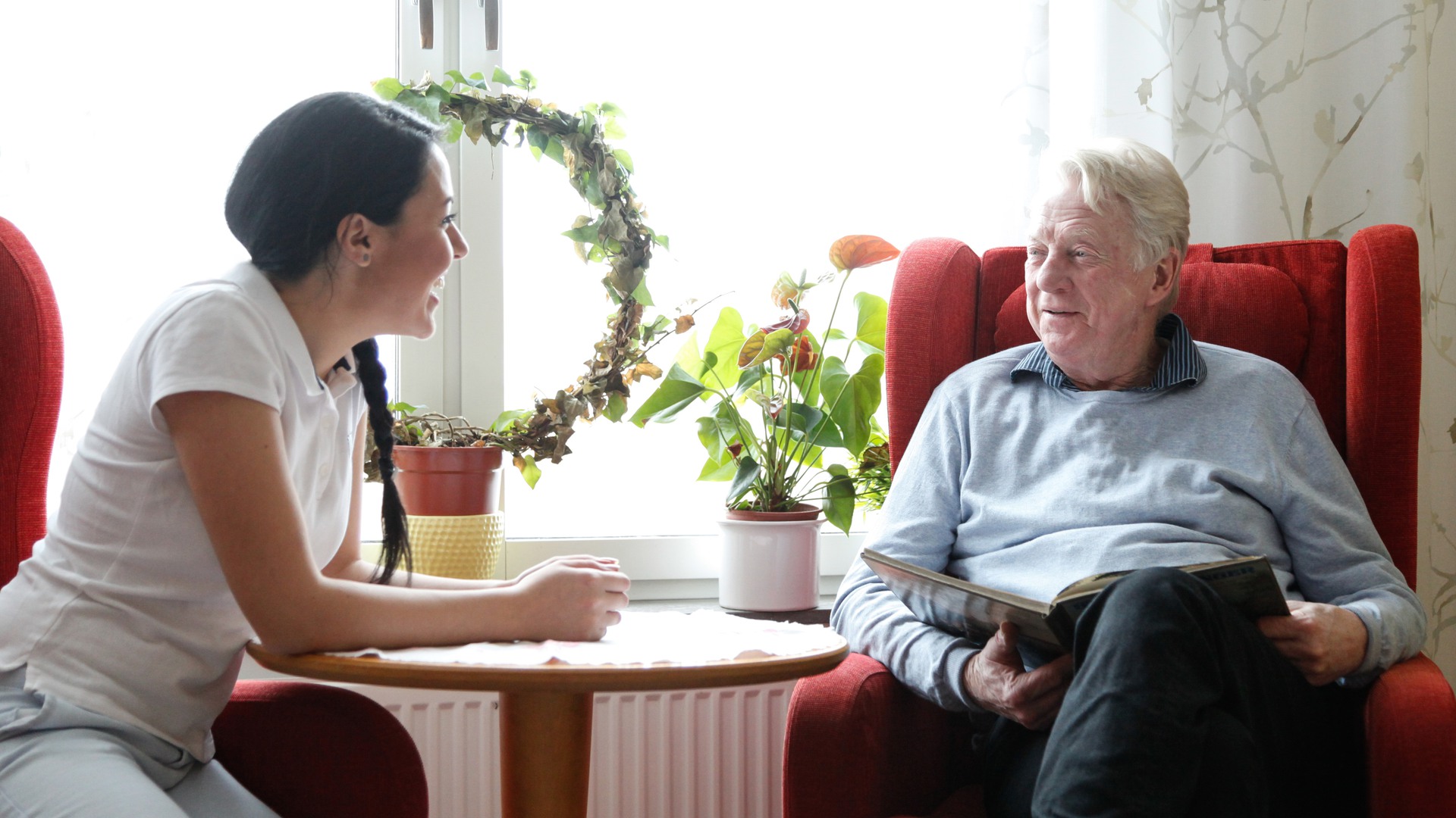 En person som jobbar i vården sitter och samtalar med en äldre man. 