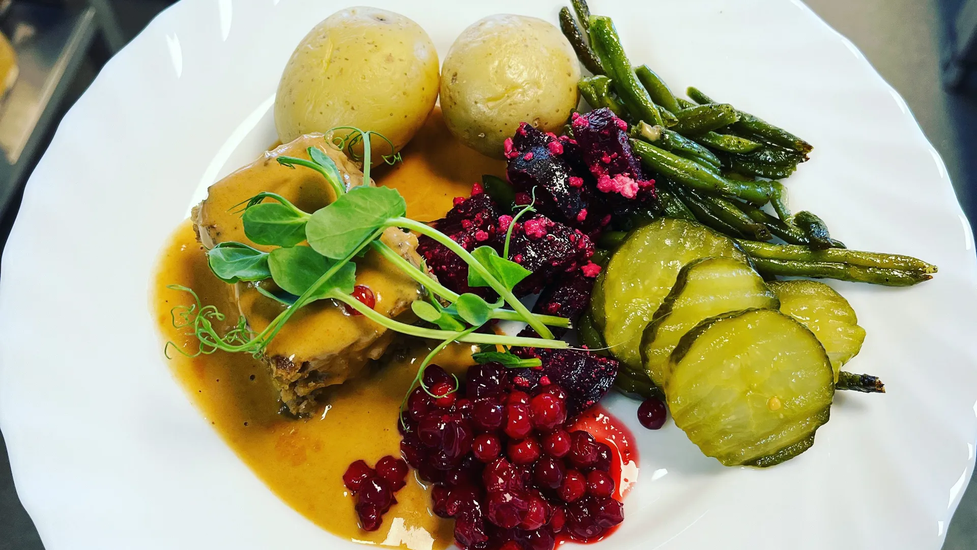 Tallrik med köttfärslimpa, potatis, rårörda lingon och grönsaker. I bakgrunden syns tillagningsköket.