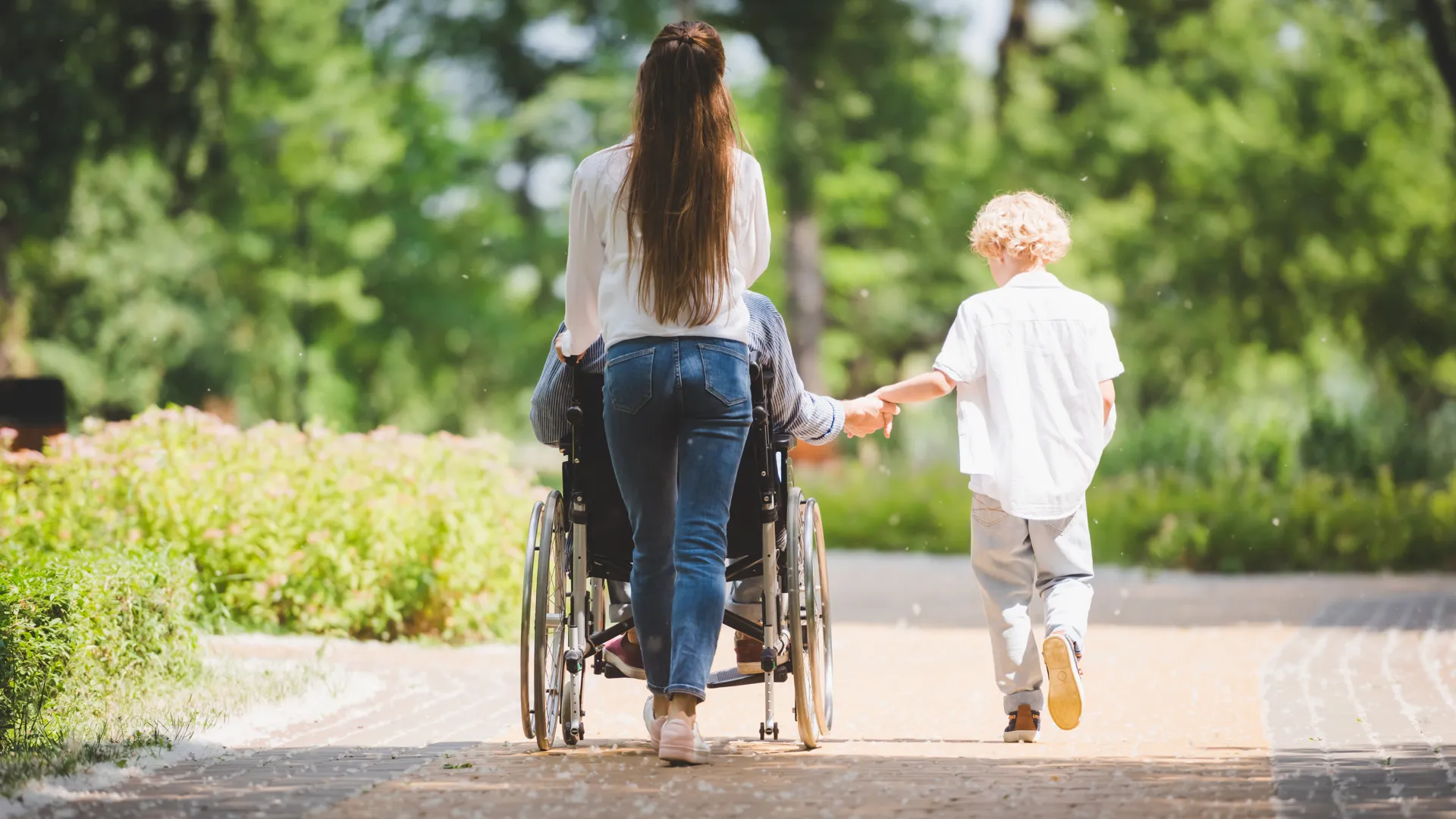 En familj på promenad i en park. Mamman skjutsar pappan som sitter i rullstol. Sonen går bredvid.