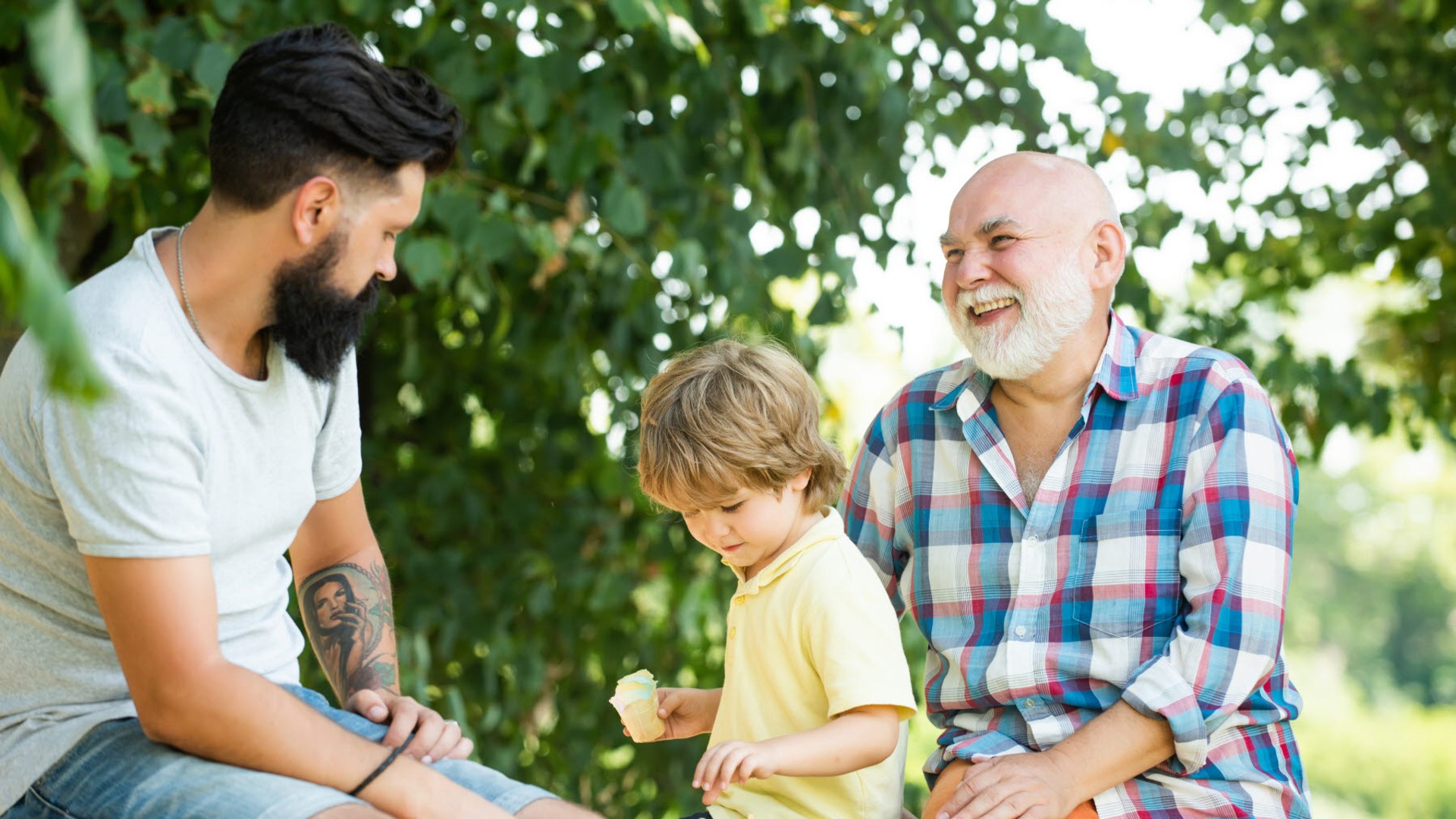 En familj med son, pappa och farförälder sitter på en bänk i solen.