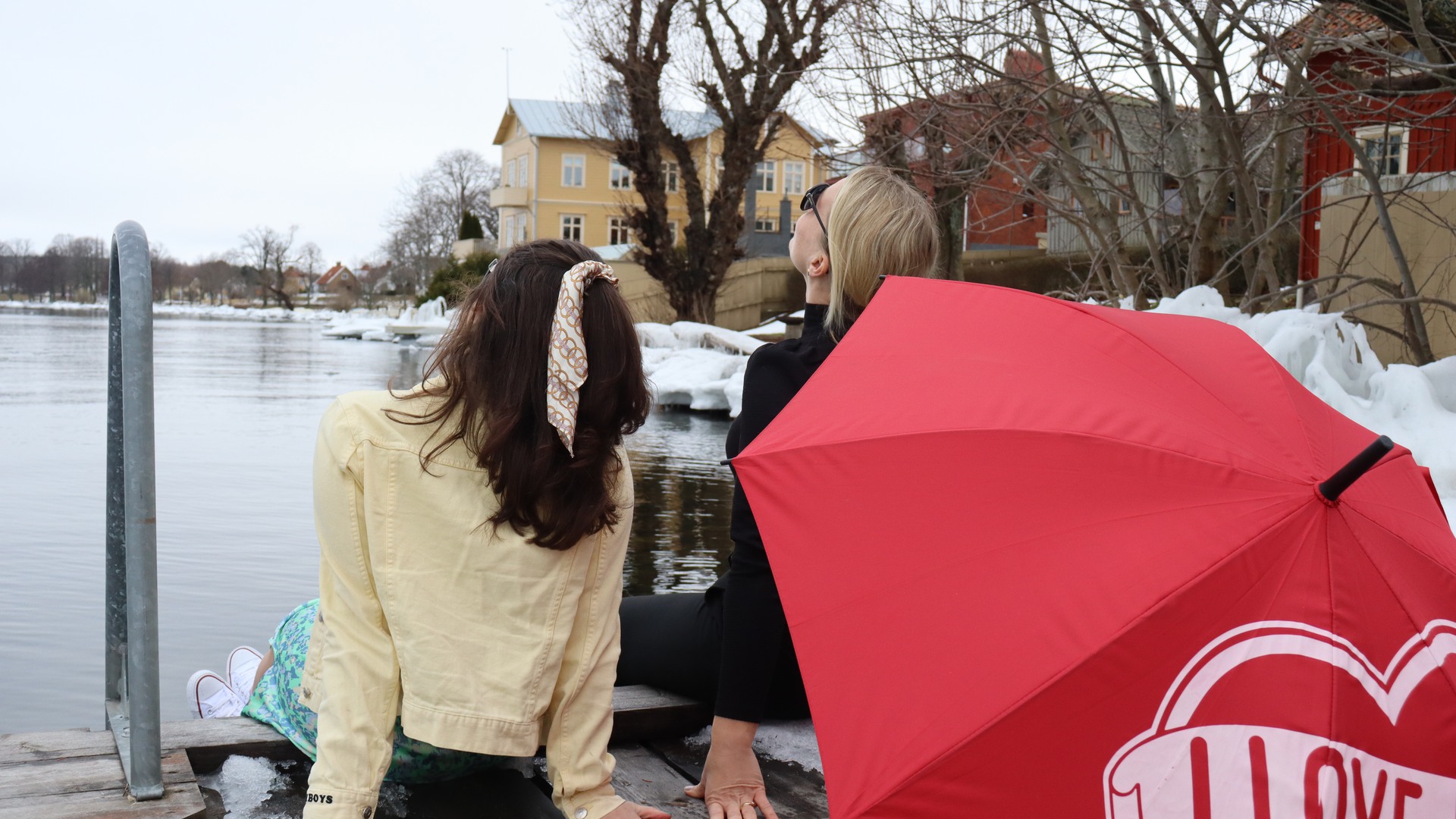 Två sommarklädda personer sitter på en brygga och njuter av ett inbillat solsken. Runt omkring dem ligger snö och träden är bara.