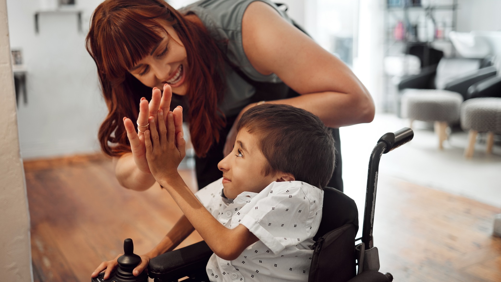 Hairdresser, high five and child in hair salon happy with hair cut or hairstyle. Disabled kid in wheelchair, stylist in salon and smile with success. Support, happiness and care for special needs boy.