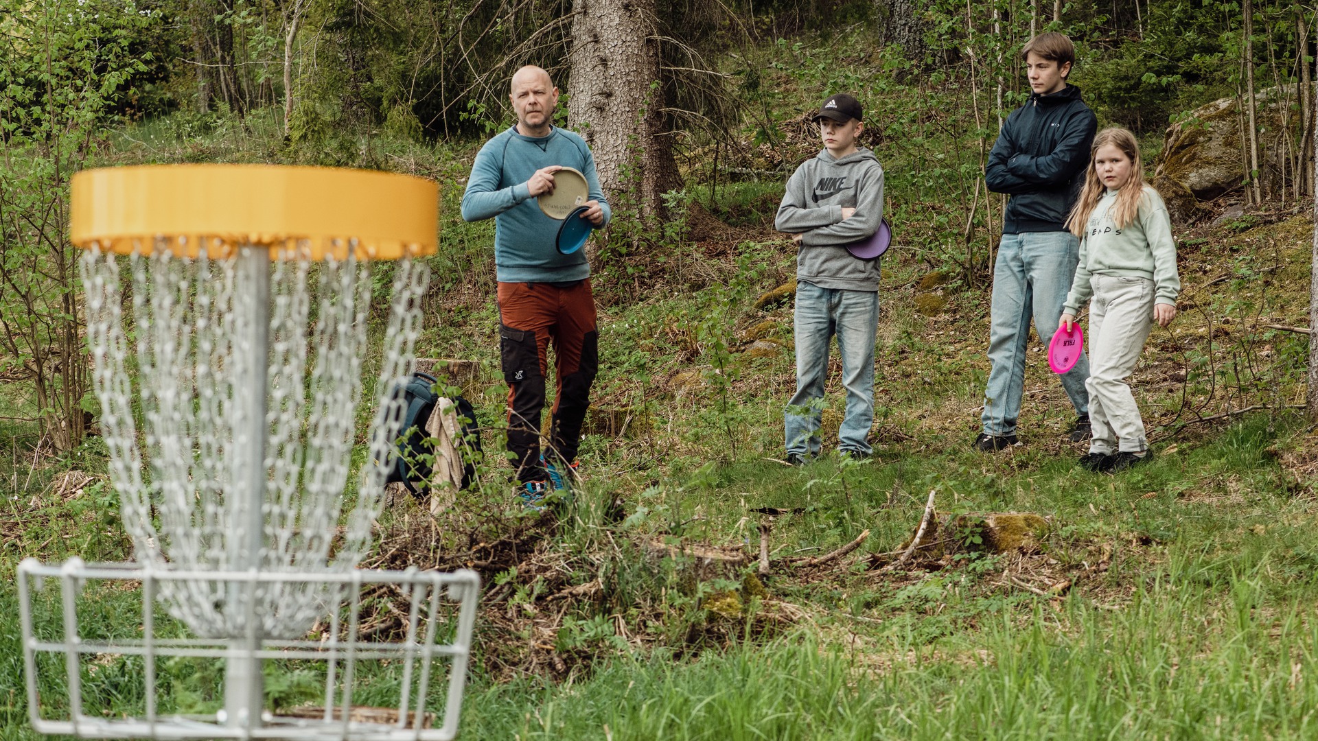 Vuxna och barn vid en korg på discgolfbanan i Högaliden.