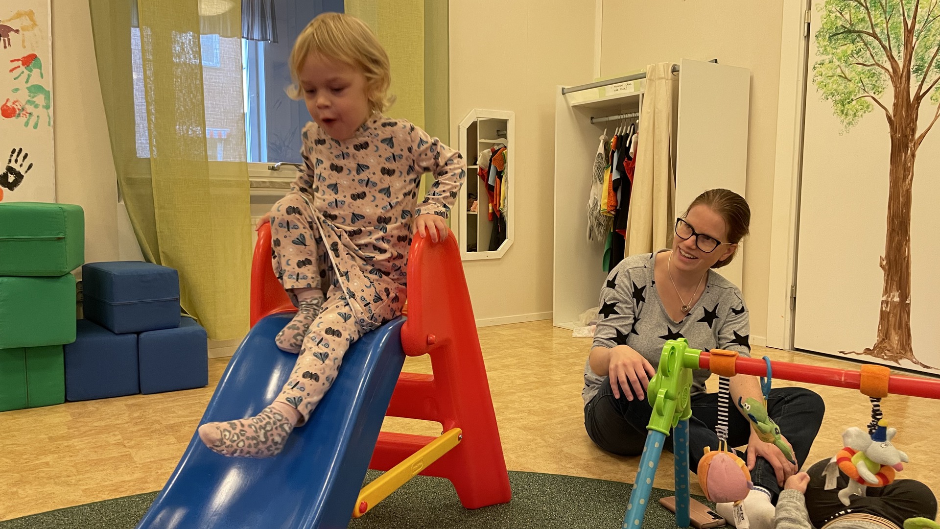 En flicka åker rutschkana inne på Öppna förskolan. Hennes mamma ser leende på henne från sidan. Framför mamman ligger en bebis i ett babygym.