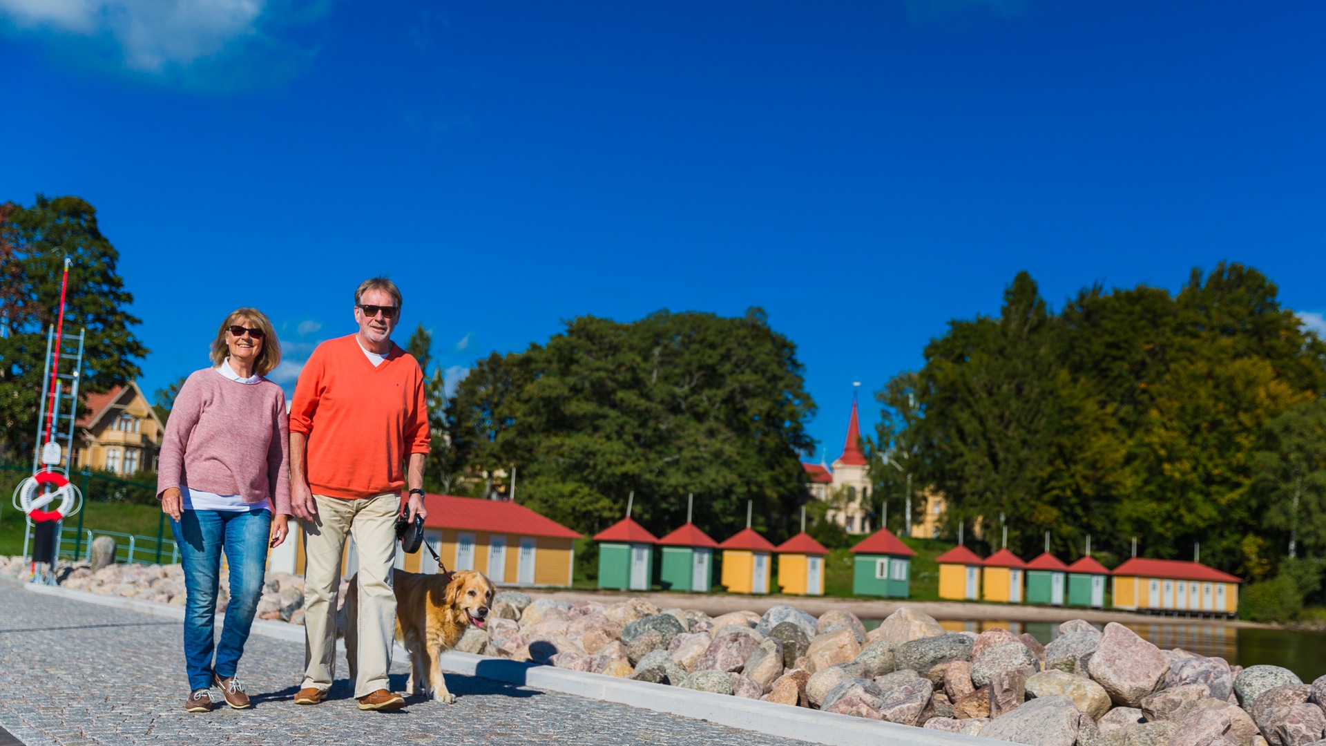 Två personer promenerarar med en hund på piren i hamnen.